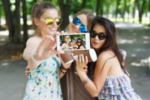 Three girls friends with coffee take away outdoors and someone POV view, taking photos at digital camera of smartphone. Young females at mobile screen, having fun in summer park. Lifestyle portrait.